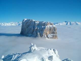 Ein schneebedecktes Bergmassiv ragt über eine dichte Wolkendecke, im Hintergrund sind weitere schneebedeckte Gipfel zu sehen. Der Himmel ist klar und blau.