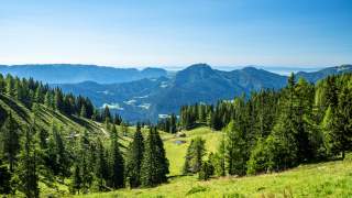 Ausblick über grüne Wiesen, Wälder, einen kleinen See und Berge am Horizont.