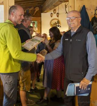 Zwei Männer schütteln sich in einem rustikalen Raum die Hände, wobei einer eine Urkunde hält und der andere ein Buch trägt
