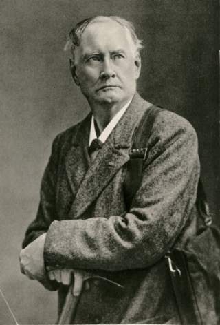 Schwarzweiß-Porträt eines älteren Mannes mit kurzem Haar in einem Mantel, der ernst blickend einen Pickel hält.