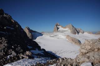 Schneebedeckte Berggipfel und Felsen unter klarem blauem Himmel in einer alpinen Landschaft. Im Vordergrund steinige, teilweise schneebedeckte Felsen.