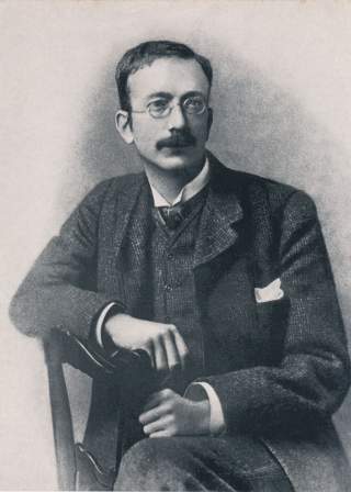 Schwarz-weiß-Porträt eines sitzenden Mannes mit Nickelbrille, Schnurrbart und Anzug, der den rechten Arm auf eine hölzerne Stuhllehne legt.