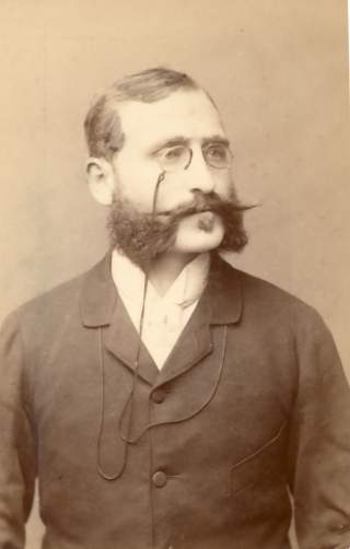 Historisches Porträt eines Mannes mit üppigem Schnurrbart, Brille mit einer Kette und dunklem Anzug vor hellem Hintergrund.