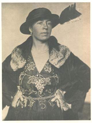 Schwarz-weiß-Porträt einer Frau in einem kunstvoll bestickten Kleid mit Pelzbesatz und Hut mit Feder, die selbstbewusst die Hände in die Seiten stemmt.