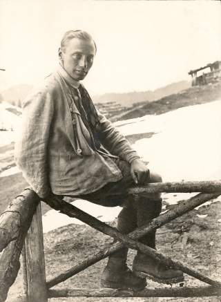 Historisches Schwarzweiß-Foto eines jungen Mannes in traditioneller Berg-Kleidung, der rücklinks auf einem rustikalen Holzzaun im alpinen Gelände sitzt. Im Hintergrund sind schneebedeckte Bergwiesen und eine Berghütte zu sehen.