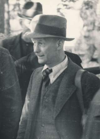 Historisches Schwarzweiß-Foto eines älteren Mannes mit Hut, Anzug und Krawatte, der im Profil nach rechts schaut, im Hintergrund sind weitere Personen unscharf zu erkennen.