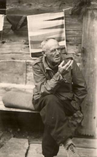 Schwarz-weiß-Foto eines älteren Mannes, der auf einer Holzbank sitzt und mit der rechten Hand gestikuliert, vor einem rustikalen Holzwand-Hintergrund.