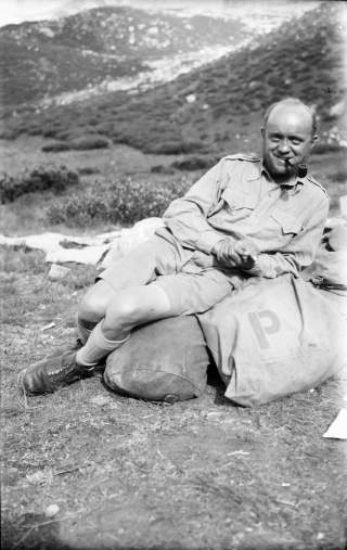 Schwarzweißfoto eines lächelnden Mannes in Felduniform mit Pfeife im Mund, der entspannt auf einem großen Sack im Freien liegt, umgeben von hügeliger Landschaft.