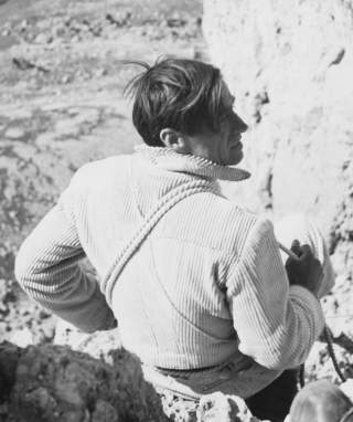 Schwarz-weiß-Foto eines Mannes in Kletterausrüstung mit Seil um den Oberkörper, der an einer Felswand sitzt oder steht und nach rechts blickt.