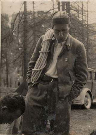 Schwarzweißfoto eines jungen Mannes mit Bergsteigerausrüstung, der eine Kletterseil-Schlinge über der Schulter trägt; im Hintergrund Wald und ein Auto.