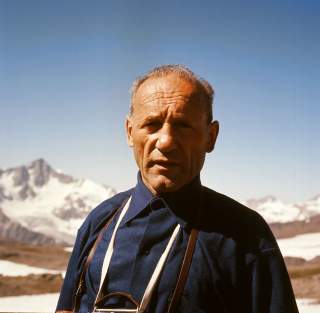 Porträt eines älteren Mannes in blauem Hemd mit Berglandschaft und Schnee im Hintergrund unter klarem Himmel.