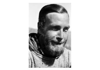 Schwarz-weiß-Porträt eines bärtigen Mannes mit zurückgekämmtem Haar, der leicht lächelnd nach rechts schaut. Er trägt einen grob gestrickten Pullover und ein Tuch.