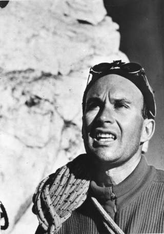 Schwarzweiß-Porträt eines Mannes mit Bergsteigerbrille auf der Stirn und Seil über der Schulter vor felsigem Hintergrund.