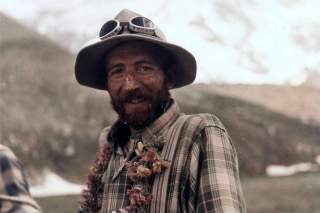 Porträt eines bärtigen Mannes mit kariertem Hemd, Hut und einer Sonnenschutzbrille für die Berge auf dem Kopf. Er trägt einen Blumenkranz um den Hals und hat die sonnenverbrannte Haut im Gesicht dick eingecremt.