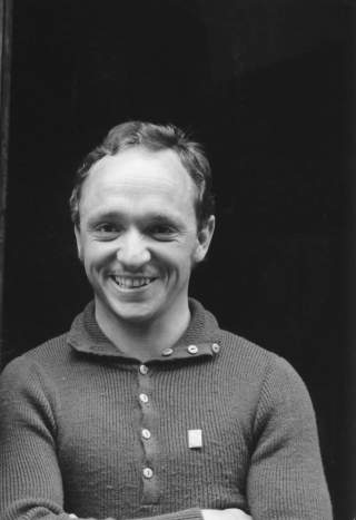 Porträt eines lächelnden Mannes in einem Pullover mit Knöpfen vor dunklem Hintergrund.