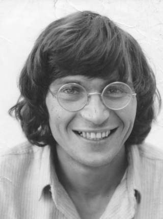 Schwarz-Weiß-Porträt eines jungen Mannes mit mittellangem Haar, der eine Brille trägt und in die Kamera lächelt.