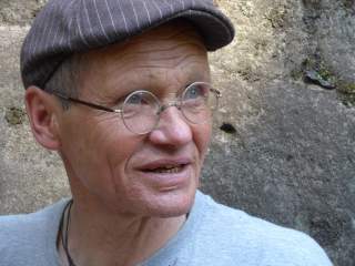 Porträt eines älteren Mannes mit Rundbrille und Schirmmütze vor einer Steinwand, der nach rechts blickt.