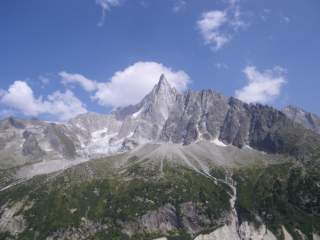 Felsige Bergspitzen mit vereinzelten Schneeflecken unter blauem Himmel mit wenigen Wolken, im Vordergrund grüne Vegetation.