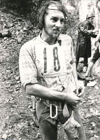 Schwarz-weiß-Foto eines jungen Mannes mit Nummernschild 18, der in Outdoor-Kletterkleidung und mit Chalkbag vor felsigem Gelände steht; im Hintergrund weitere Personen mit Kletterausrüstung und Regenjacke.