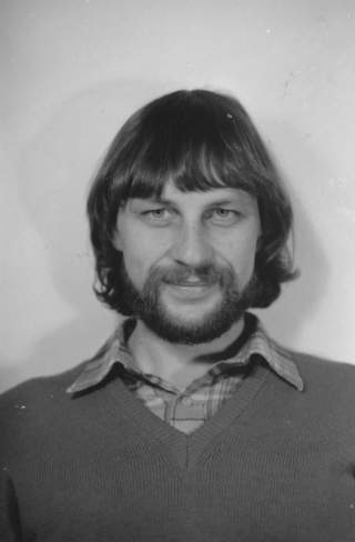 Schwarz-weiß-Porträt eines Mannes mit mittellangen Haaren, Vollbart und kariertem Hemd unter einem Pullover, der in die Kamera lächelt.