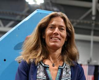 Frau mit langem, leicht gewelltem Haar, die lächelnd in die Kamera schaut, vor einem blauen Objekt und unscharfem Hintergrund mit amerikanischer Flagge.