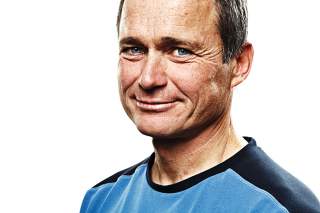 Porträt eines lächelnden Mannes mit kurzen braunen Haaren und blauen Augen, der ein blau-schwarzes Oberteil trägt, vor weißem Hintergrund.