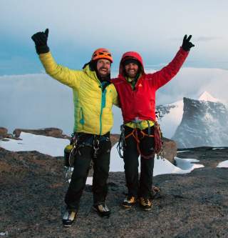 Zwei bergsteigende Männer in warmer, farbiger Ausrüstung und Helm posieren glücklich auf einem felsigen, verschneiten Gipfel mit Berglandschaft im Hintergrund.