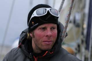 Porträt eines Mannes mit rotem Gesicht, der eine schwarze Mütze mit weißer Sonnenbrille und eine graue Jacke trägt, im nicht-fokussierten Hintergrund Kletterseile