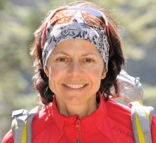 Porträt einer lächelnden Frau mit roter Outdoorjacke, Stirnband und Sonnenbrille, die einen Rucksack trägt, vor unscharfem Waldhintergrund.