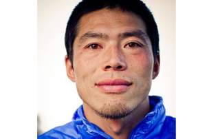 Porträt eines jungen Mannes mit kurzem schwarzen Haar und leichtem Bart, der in einer blauen Jacke direkt in die Kamera blickt.