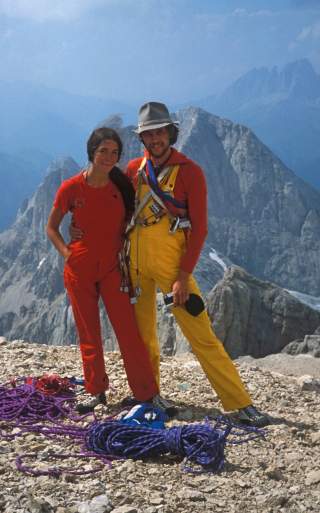Zwei Bergsteiger in roter und gelber Kletterkleidung posieren umarmt auf einem felsigen Gipfel vor einer Bergkulisse mit gesammelten Kletterseilen am Boden.