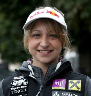 Porträt einer lächelnden Frau mit weißem Red Bull-Visor und schwarzer Sportjacke mit verschiedenen Sponsorenlogos vor unscharfem Hintergrund.