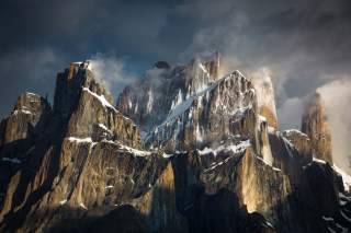 Felsige, schroffe Berggipfel mit Schneeflecken, umgeben von Nebel und dramatisch beleuchtetem Himmel.