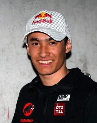 Porträt eines lächelnden jungen Mannes mit schwarzer Jacke und kariertem Red Bull-Basecap vor grauer Wand.