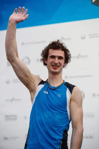 Junger Mann in blauem Sportshirt hebt lächelnd die rechte Hand zum Winken.