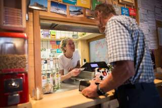 Am Tresenfenster einer Berghütte sind eine Frau und ein älterer Mann im Gespräch. Im Hintergrund sind verschiedene Produkte und bunte Karten an der Wand zu sehen.