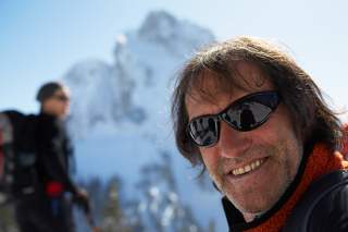 Mann mit Sonnenbrille lächelt in die Kamera, im Hintergrund schneebedeckter Berg und eine weitere Person.