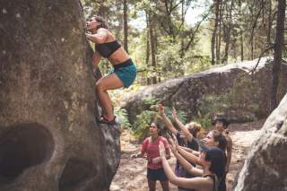 Mensch bouldert am Fels, eine Gruppe steht am Boden zum Spotten.