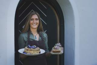 Küchenchefin Evelyn der Franz-Fischer-Hütte hält frischen Kuchen und Brot in der Hand und steht vor neutralem Hintergrund