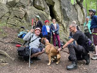Vor einer Felswand kniet ein Mann mit Blindenstock streichelt einen Hund und hat eine Kameratasche umgehängt.