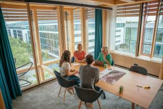 Vier Personen sitzen in einem modernen, lichtdurchfluteten Besprechungsraum an einem Holztisch und führen eine Diskussion, umgeben von großen Fenstern mit Blick auf ein Bürogebäude.