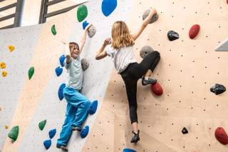 Zwei Kinder klettern an einer Indoor-Boulderwand mit bunten Griffen, gerade wollen sie freudig abklatschen.