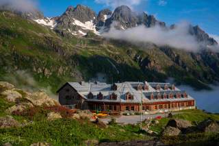 Berghütte in einer grünen Alpenlandschaft, im Hintergrund schroffe, teils schneebedeckte Berggipfel und tiefe Wolken.