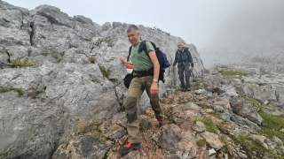 Zwei Männer wandern auf einem felsigen Gebirgspfad. Es ist neblig und felsig mit spärlicher Vegetation.