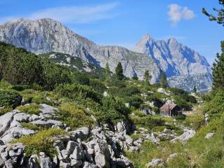 Berglandschaft mit steinigen Hängen, grünen Büschen und Bäumen, im Hintergrund stehen markante Felsgipfel unter blauem Himmel, eine kleine Holzhütte ist am rechten Bildrand zu sehen.