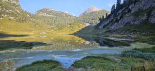 Bergsee mit spiegelglatter Wasseroberfläche, umgeben von bewaldeten und felsigen Bergen, im Hintergrund eine Berghütte unter blauem Himmel.