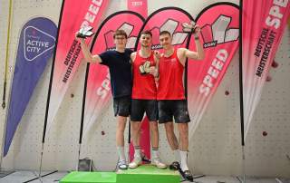 Drei junge Männer in Sportbekleidung stehen auf einem Siegerpodest, halten Pokale hoch und posieren vor Fahnen mit der Aufschrift „Deutsche Meisterschaft Speed“.