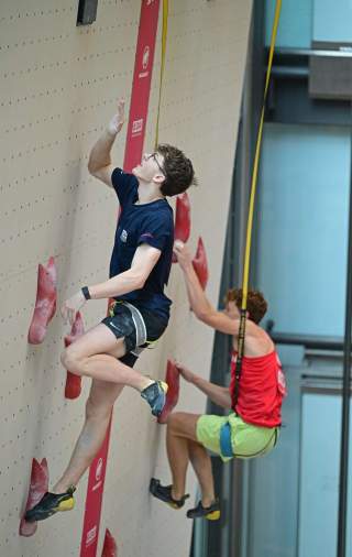 Zwei junge Männer klettern an einer Kletterwand mit roten Griffen, einer trägt ein blaues T-Shirt und der andere ein rotes. Beide sind mit Klettersicherungsgurten ausgestattet.