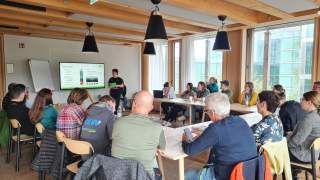 Gruppe von Menschen sitzt in einem modernen, hellen Seminarraum und hört einer Person zu, die vorne an einem Bildschirm präsentiert.