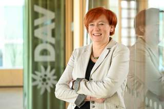 Portrait einer lächelnden Frau mit roten Haaren und einer beigen Bluse die an einer Glaswand lehnt. Im Hintergrund ist das DAV Logo zu finden.